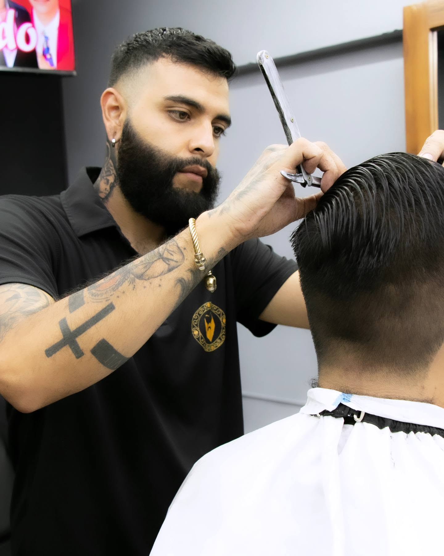 Barbero con tatuajes en el brazo repasando con navaja el contorno del cabello de un cliente, letrero de neón al fondo