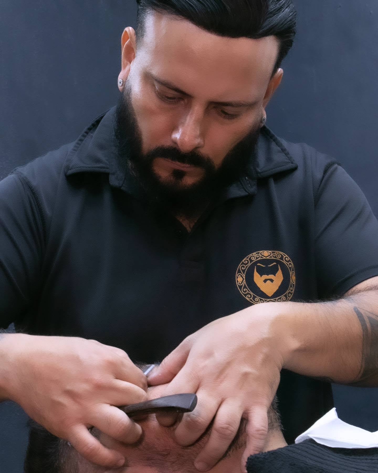 Barbero de Mr. Pedro, con polo negro y el emblema dorado bordado, perfilando la barba de un cliente con navaja