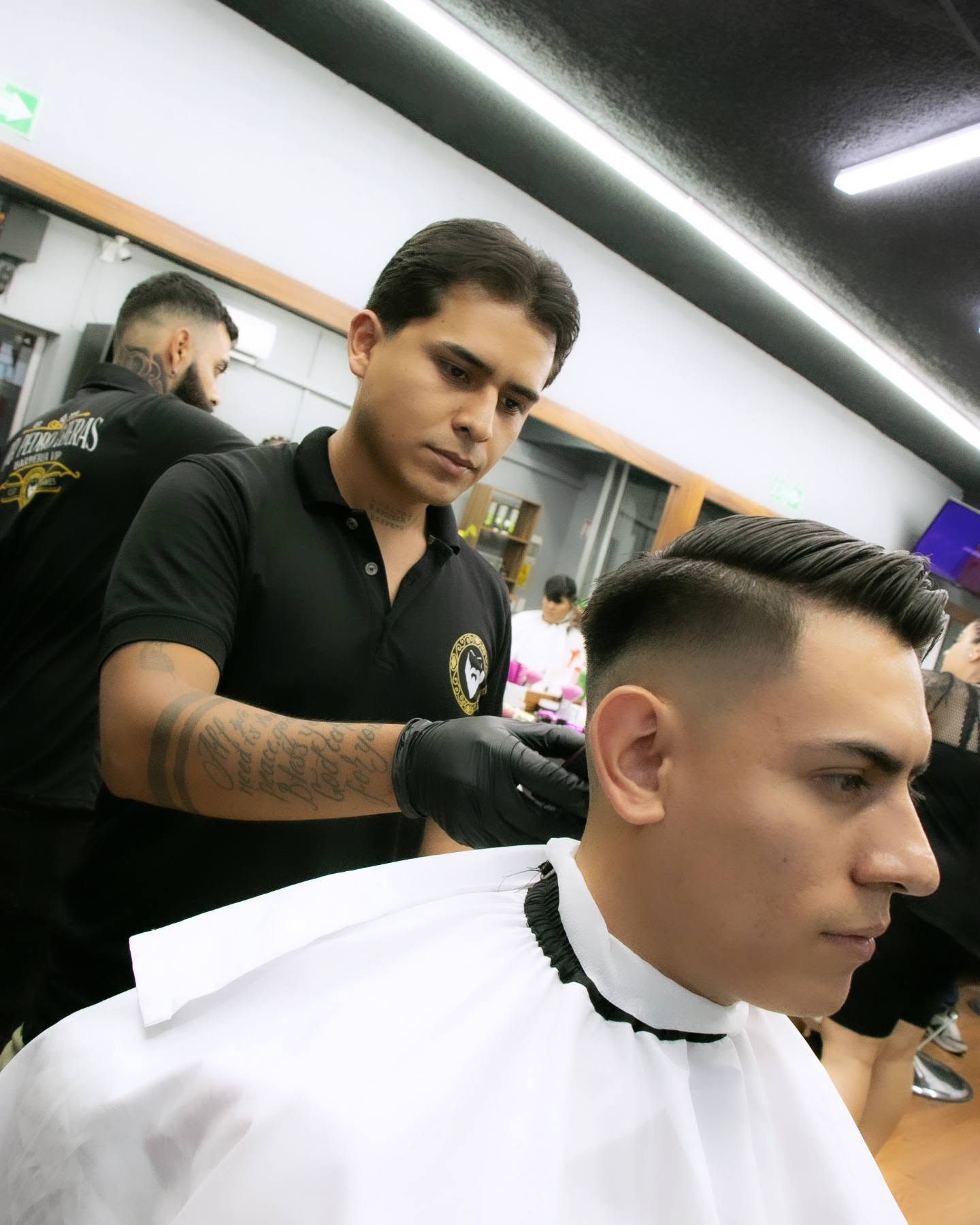 Barbero con guantes negros marcando un fade con máquina en la nuca de un cliente, con otro barbero trabajando al fondo
