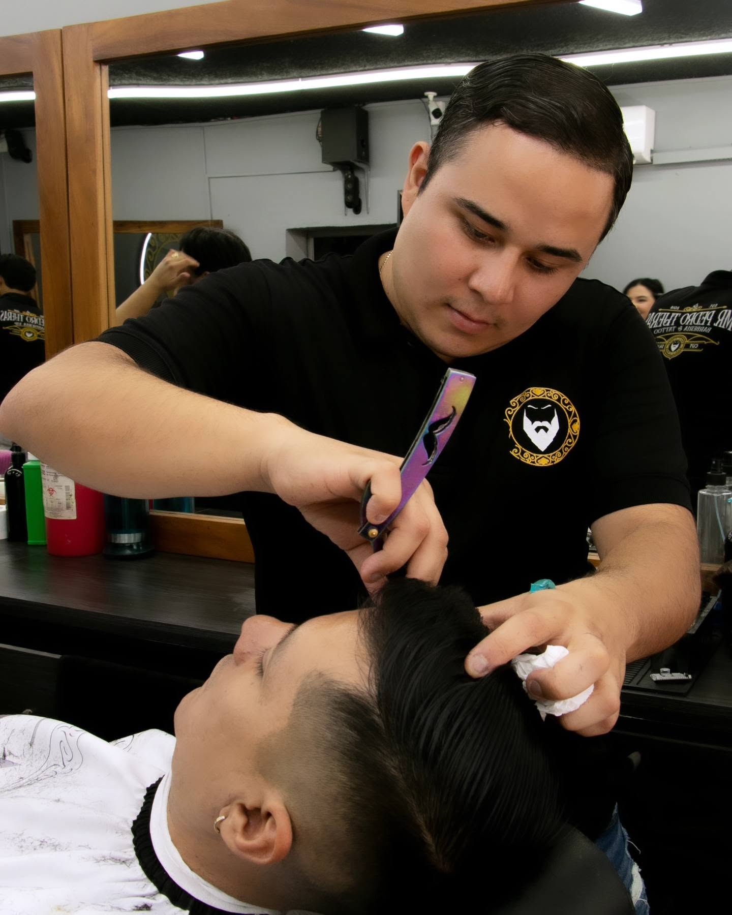 Barbero perfilando con navaja la línea del cabello de un cliente recostado, frente a los espejos de madera de la barbería