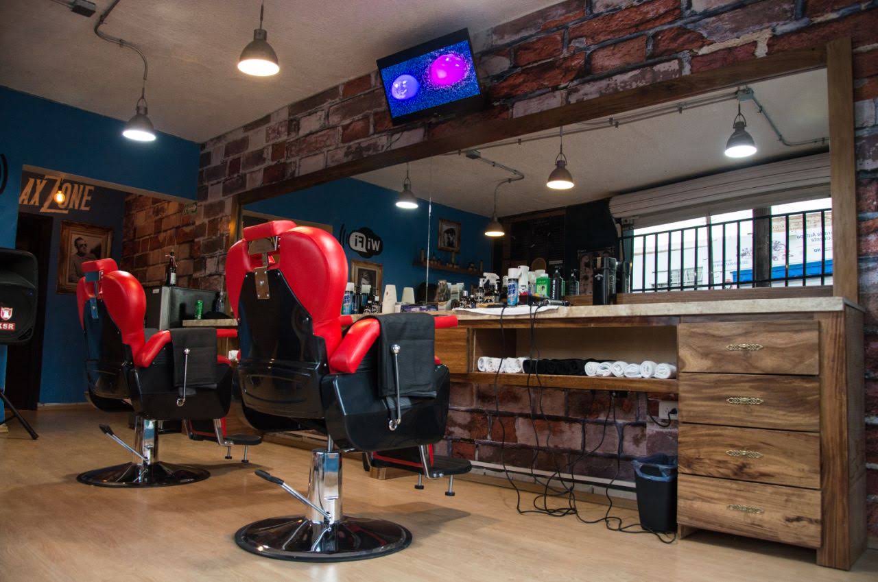Interior de la barbería con tres sillones rojos y negros, muro de ladrillo expuesto, barra de madera con productos y toallas enrolladas